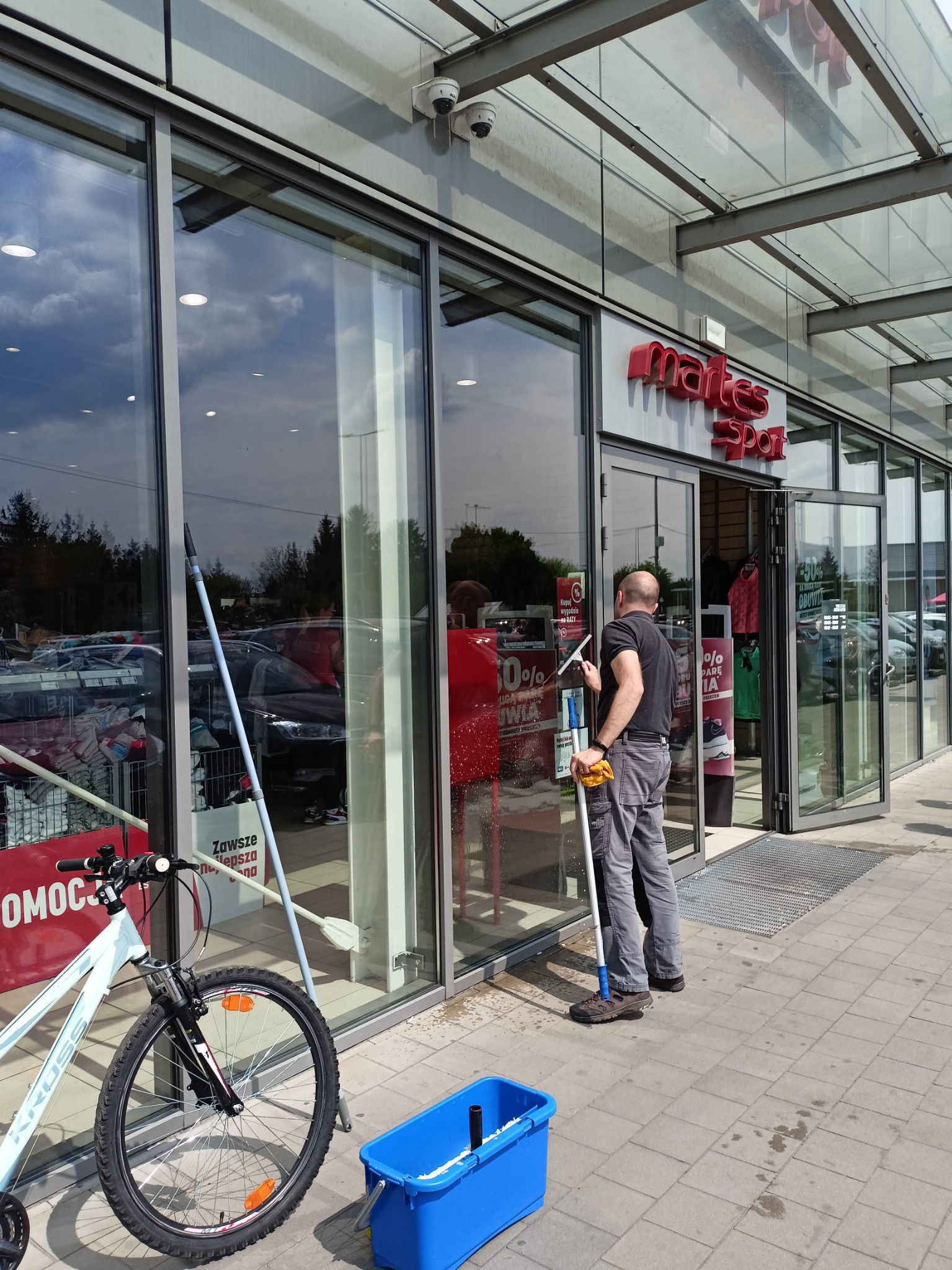 Mężczyzna w szarych spodniach i czarnej koszulce czyści szklaną witrynę sklepu sportowego za pomocą ściągaczki, obok stoi niebieskie wiadro z płynem i rower.