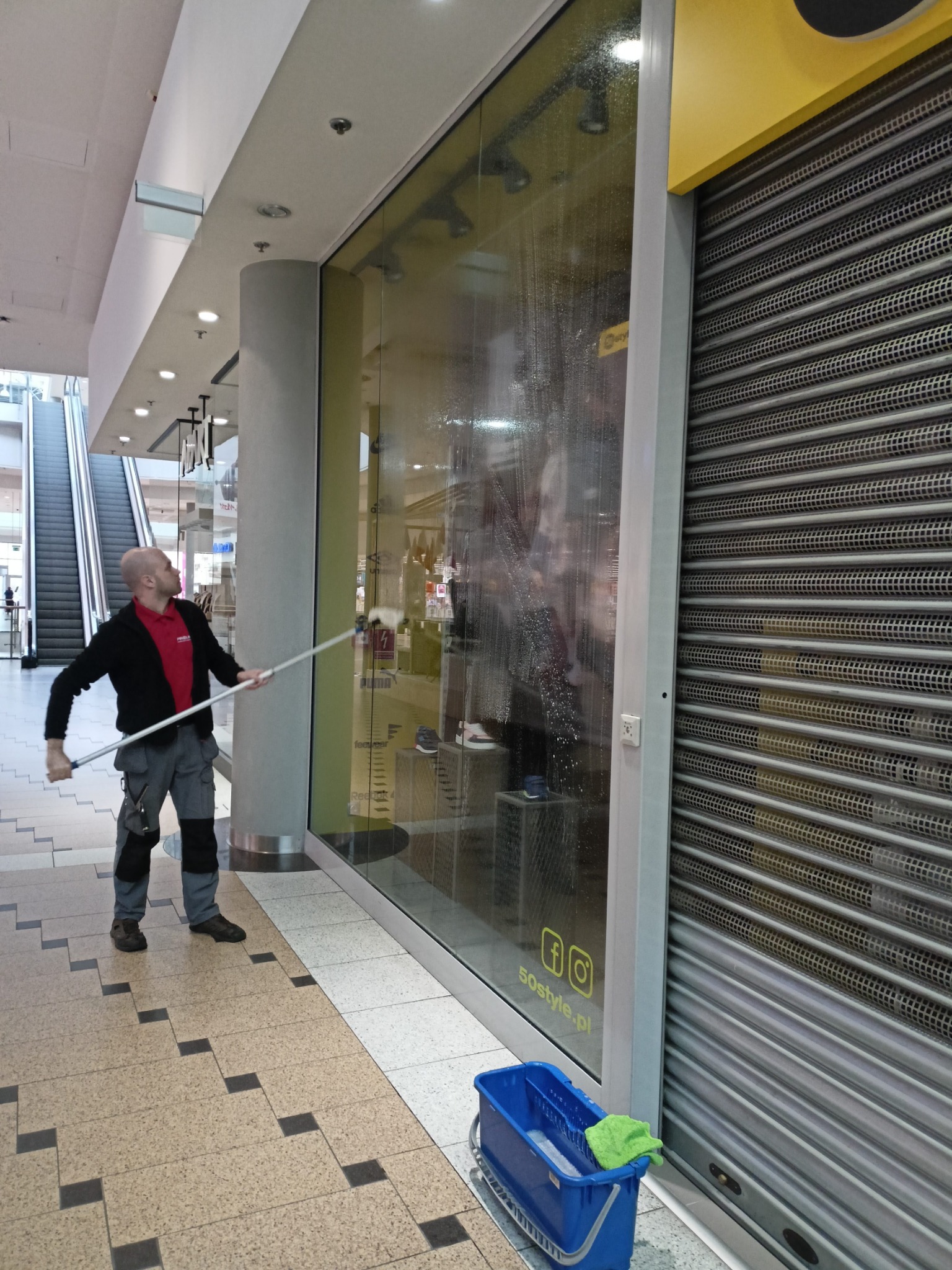 Pracownik myje okno wystawowe sklepu w galerii handlowej za pomocą mopa teleskopowego, obok zwinięta roleta i niebieskie wiadro z płynem.