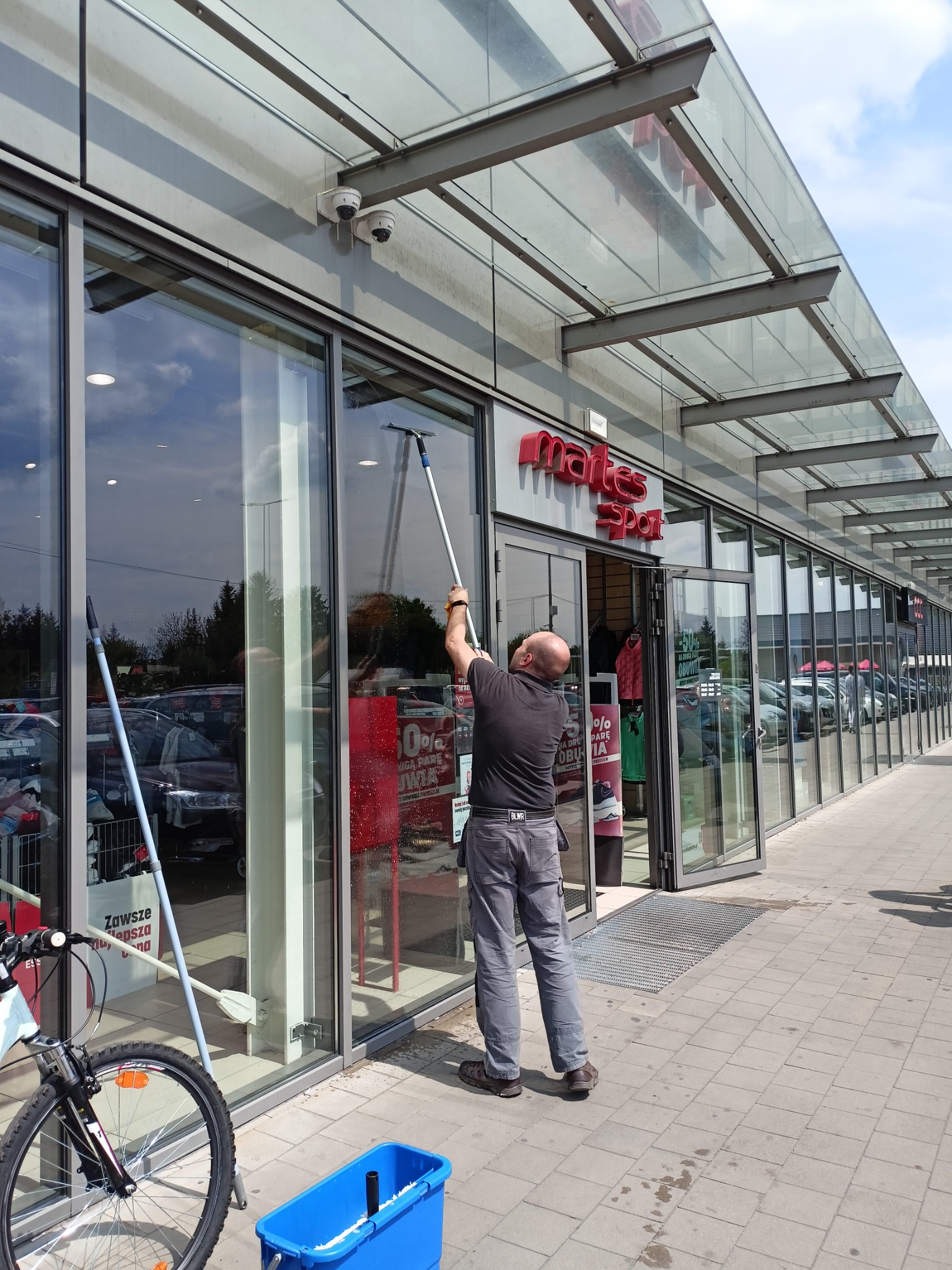 Mężczyzna myje okna wystawowe sklepu za pomocą długiego uchwytu z myjką, widoczny rower i niebieskie wiadro z płynem, sklep 'martes sport' w tle, widok z zewnątrz.