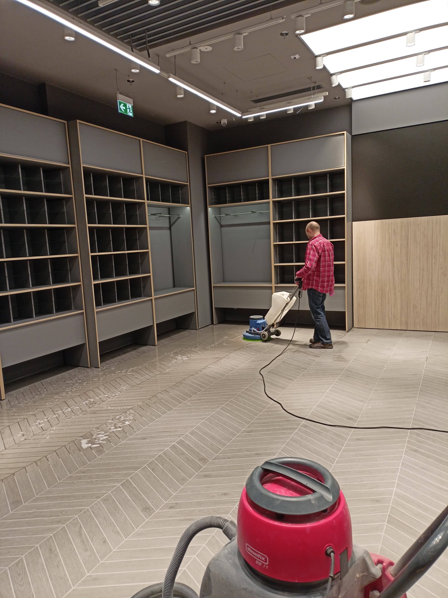 Mężczyzna w czerwonej koszuli flanelowej czyści podłogę w lokalu komercyjnym za pomocą maszyny do czyszczenia podłóg, widoczne puste regały i ślady wilgoci na podłodze ułożonej w jodełkę.