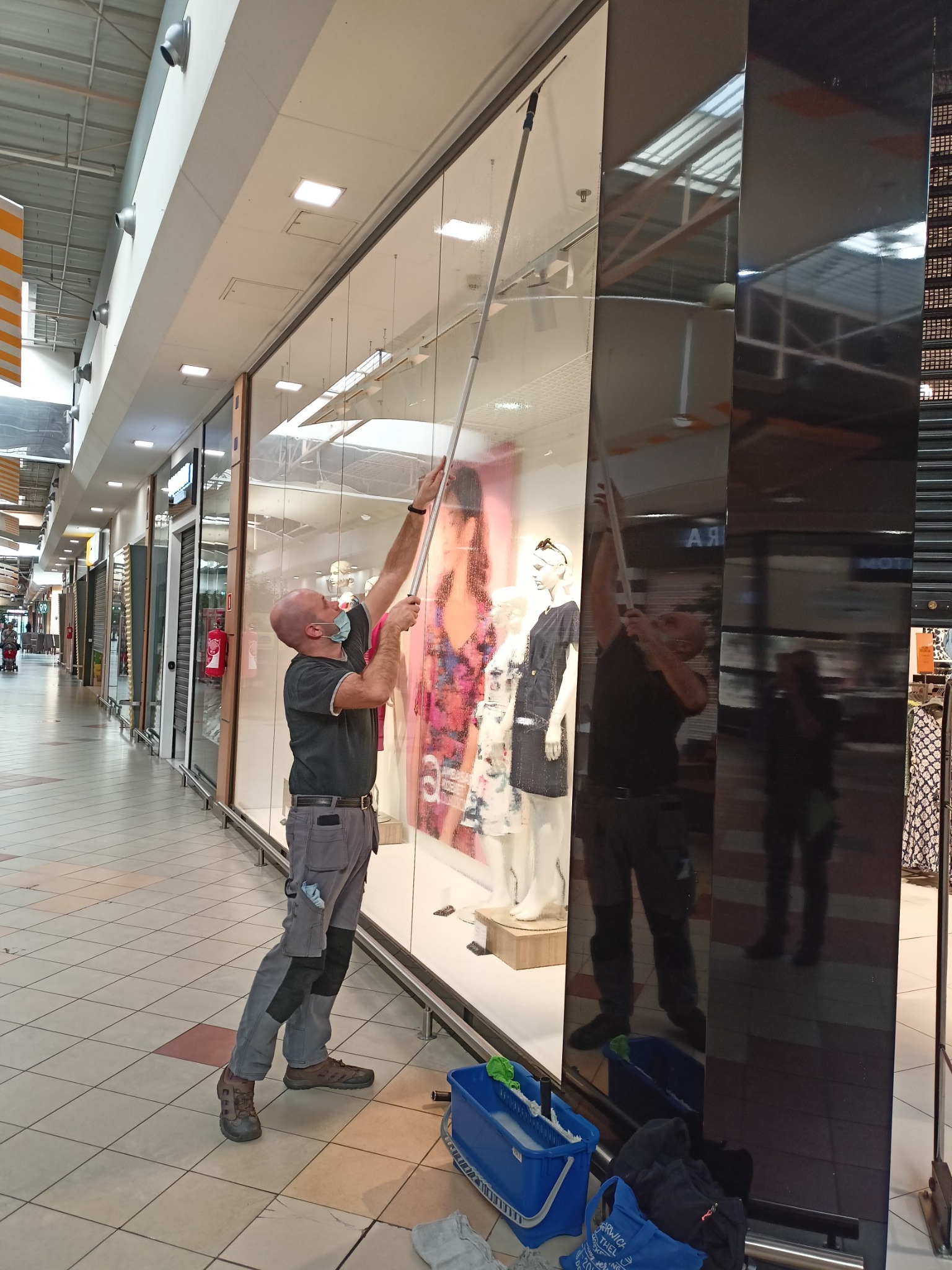 Pracownik w masce, w szarych spodniach roboczych, czyści witrynę sklepową w galerii handlowej za pomocą mopa teleskopowego; widoczne niebieskie wiadro i torba.