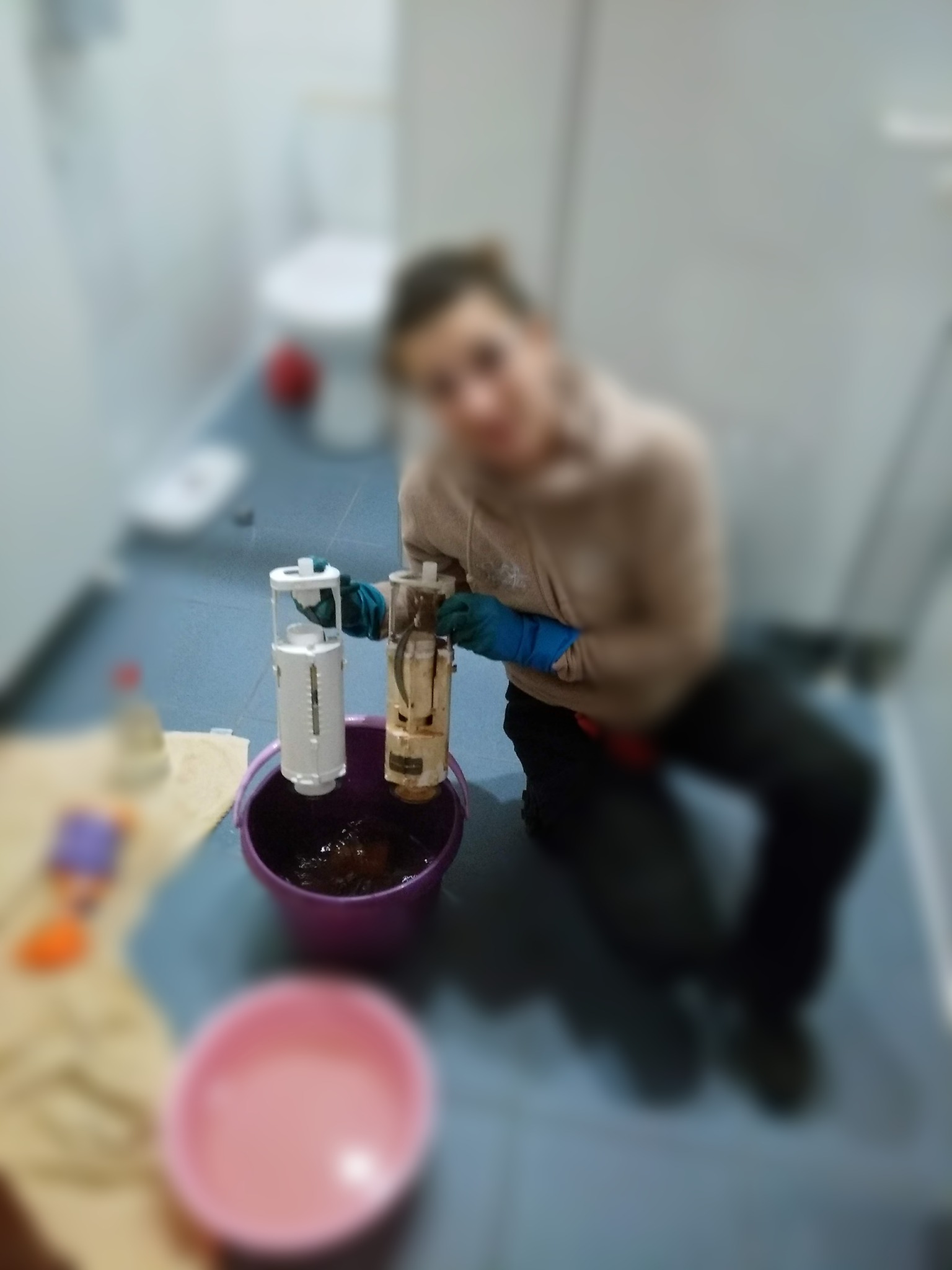 Osoba w rękawicach trzymająca dwa elementy spłuczki toaletowej nad fioletowym wiadrem, w tle widoczna toaleta i podłoga w łazience.