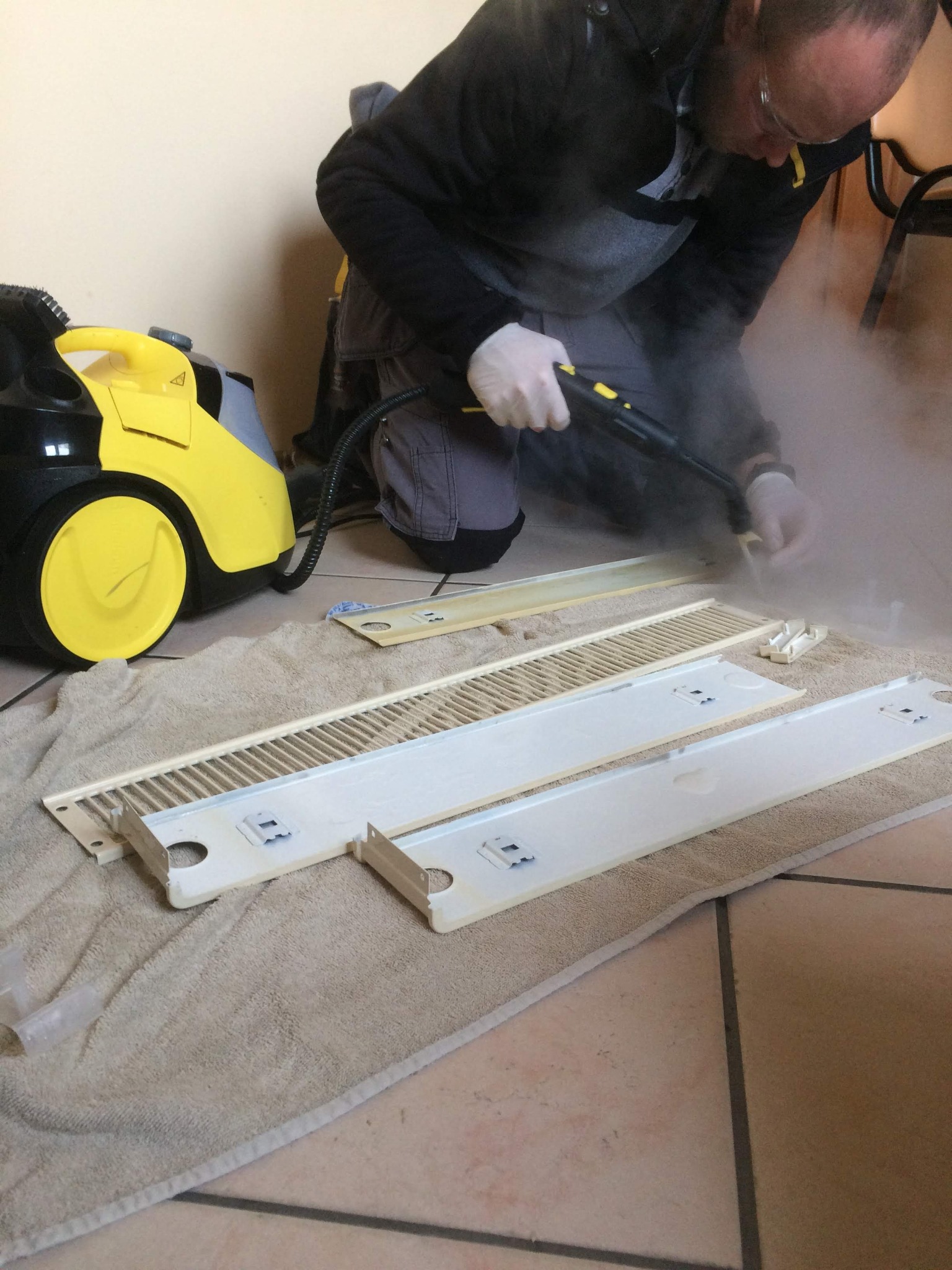 Czyszczenie parowe elementów grzejnika przez osobę w rękawicach ochronnych, na podłodze leży beżowy ręcznik, obok żółto-czarny odkurzacz parowy.