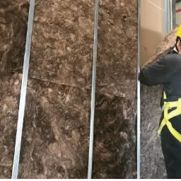 Montaż izolacji z wełny mineralnej między metalowymi profilami rusztu pod płytę gipsowo-kartonową, pracownik w żółtym kasku i uprzęży zabezpieczającej.