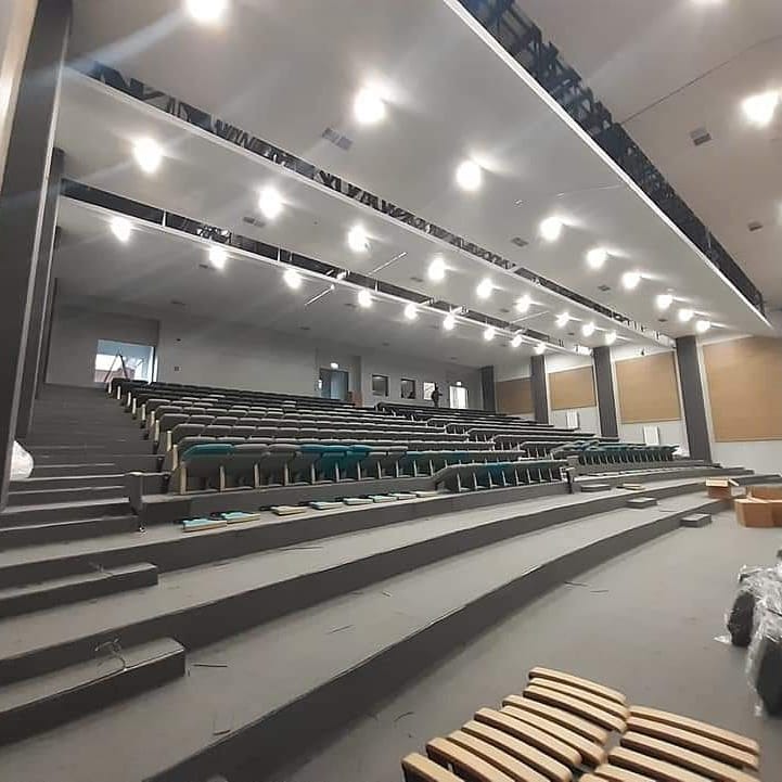 Sala konferencyjna z rzędami krzeseł na stopniach, pomalowanymi szarymi elementami i nowoczesnym oświetleniem sufitowym, w trakcie prac wykończeniowych.