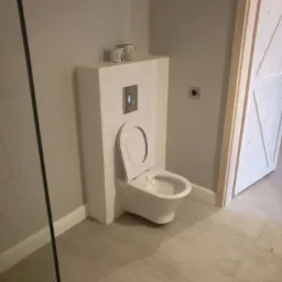 Nowoczesna toaleta podwieszana w obudowie z białych płytek, z dwoma kubkami na górze, zamontowana w szarej łazience z jasną podłogą i drzwiami.