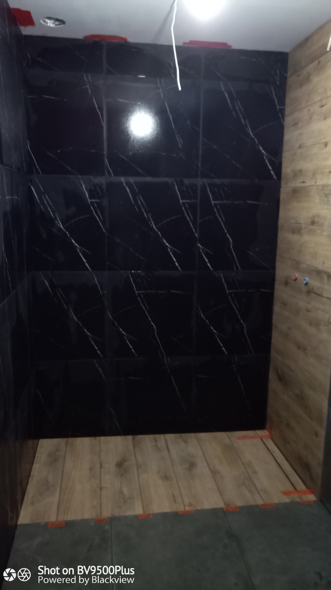 Wnętrze łazienki w trakcie remontu: ściany wyłożone czarnymi płytkami z białym wzorem marmuru, drewnopodobne panele na ścianie, podłoga z paneli w trakcie układania, widoczne krzyżyki dystansowe.