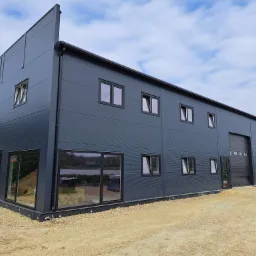 Nowoczesna hala z szarej blachy trapezowej z oknami i bramą segmentową. Widok z zewnątrz na plac budowy, błękitne niebo z chmurami w tle.