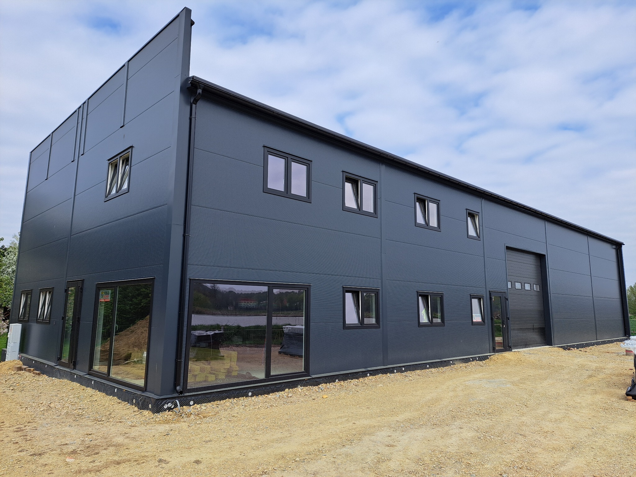 Nowoczesna hala z szarej blachy trapezowej z oknami i bramą segmentową. Widok z zewnątrz na plac budowy, błękitne niebo z chmurami w tle.