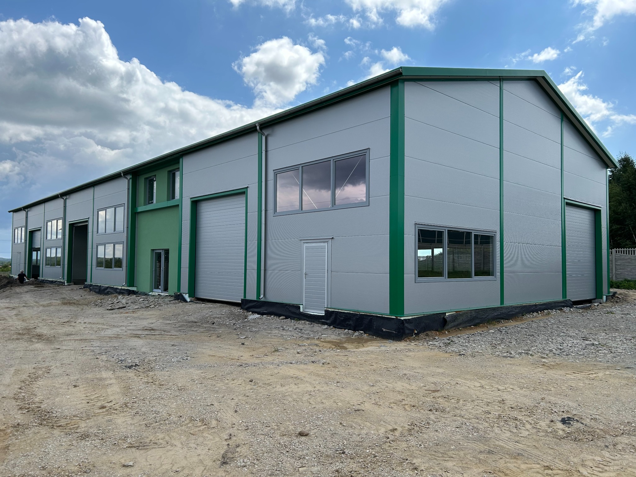 Nowoczesna hala magazynowa z szarej blachy trapezowej z zielonymi akcentami. Widoczne okna, brama garażowa i drzwi wejściowe. Na zewnątrz teren przygotowany pod utwardzenie.