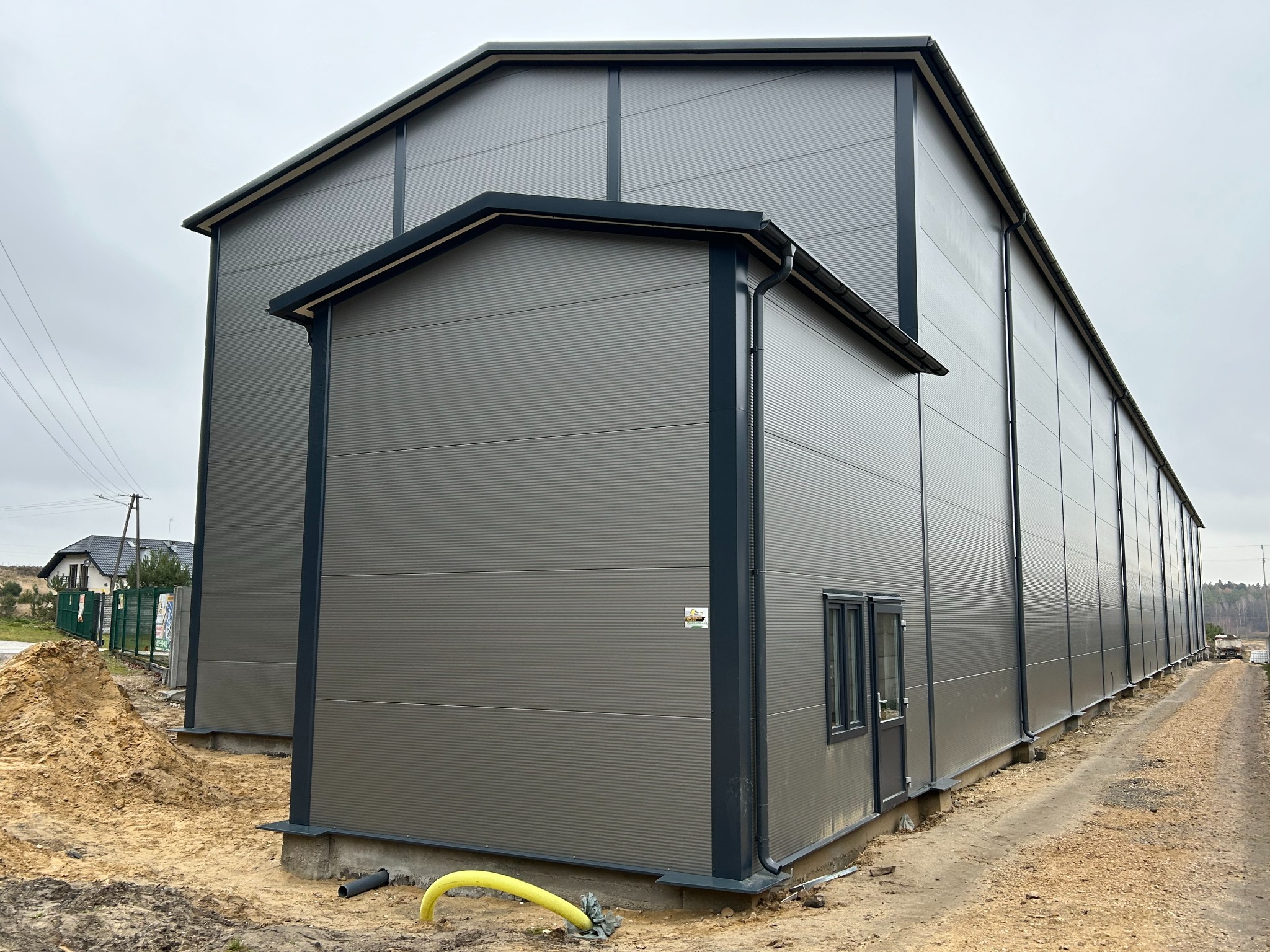 Szara hala magazynowa z dobudówką i widoczną rurą na zewnątrz. Budynek z blachy trapezowej, z ciemnymi obróbkami blacharskimi. Na drugim planie dom.