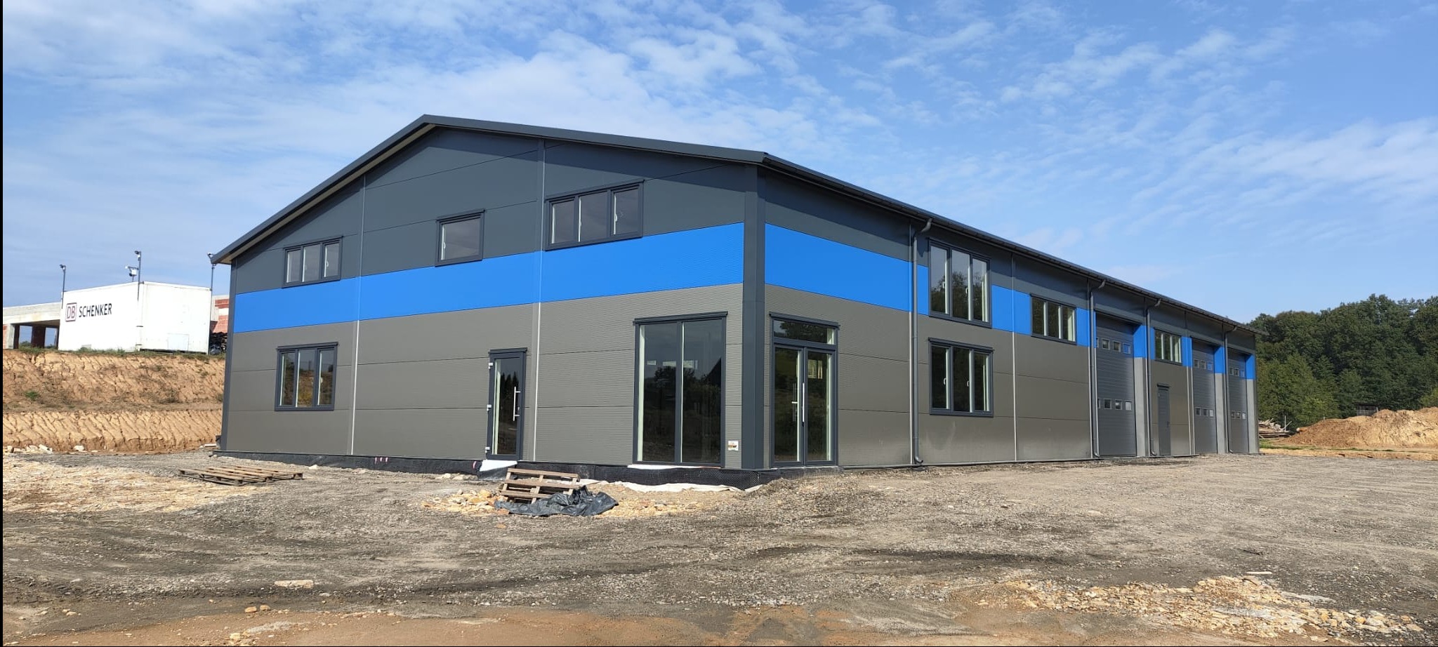 Nowoczesna hala magazynowa w szaro-niebieskiej kolorystyce, z widocznymi oknami i bramami, na tle błękitnego nieba i zieleni drzew. Widoczne palety przed budynkiem.