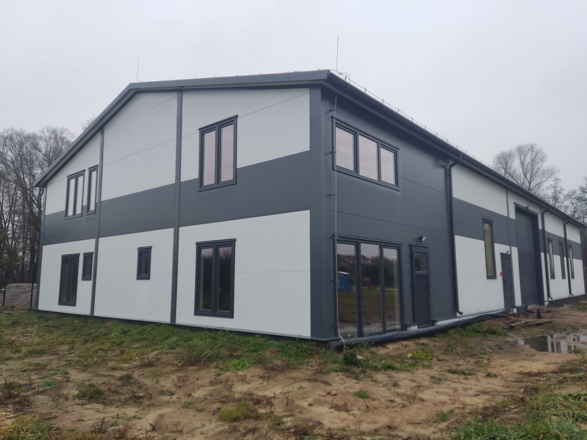 Nowoczesna hala stalowa z szaro-białą elewacją, widoczne okna i drzwi. Budynek w otoczeniu zieleni i ziemi, pochmurne niebo w tle. Ukończona realizacja.