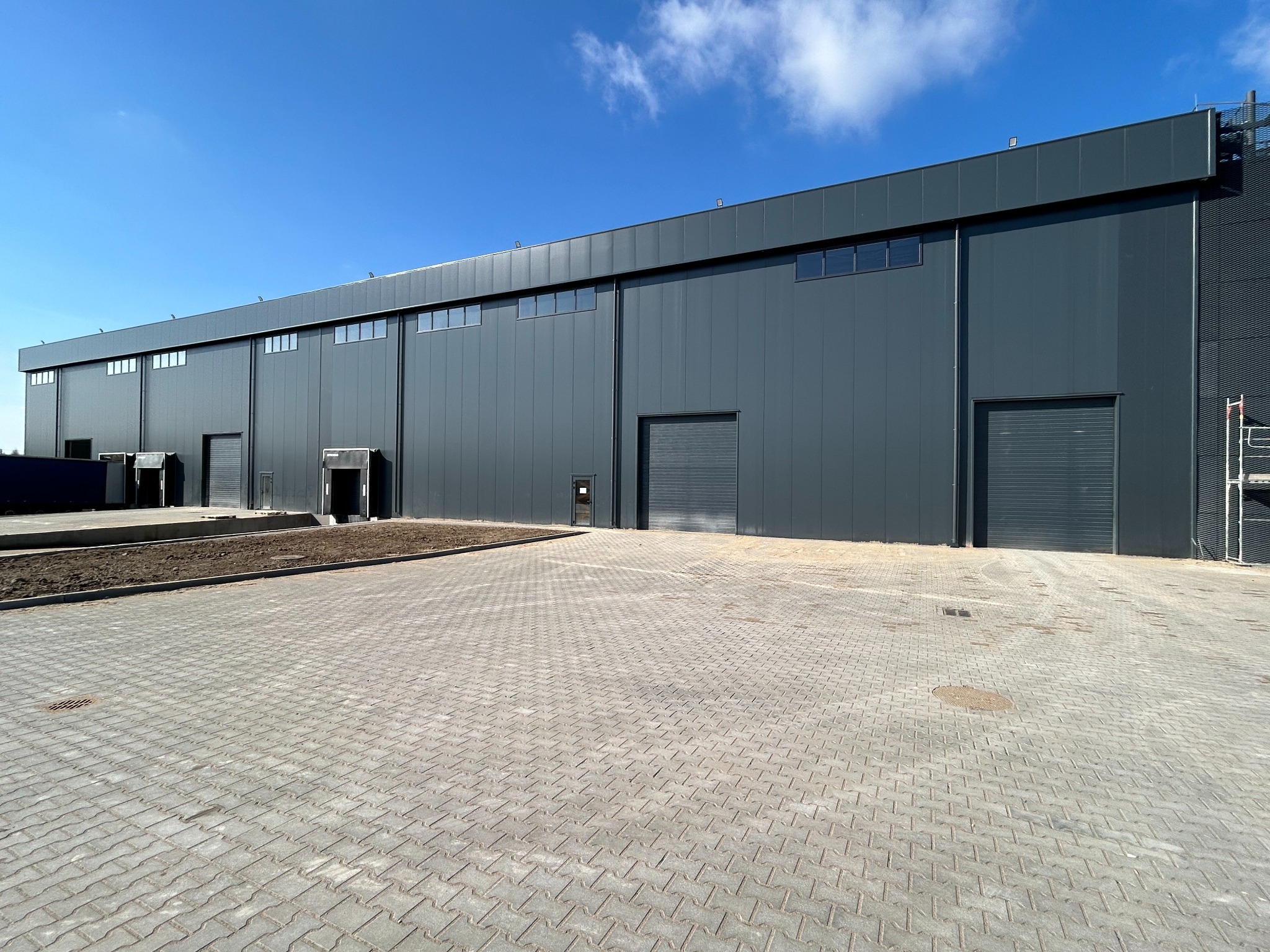 Nowoczesna hala magazynowa z szarej blachy trapezowej, widok z zewnątrz, błękitne niebo, kostka brukowa. Widoczne bramy wjazdowe i okna.