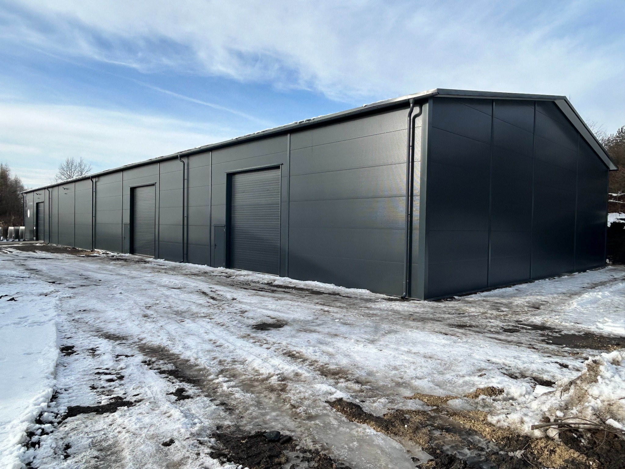 Nowoczesna, szara hala z blachy trapezowej z trzema bramami rolowanymi, na tle zimowego krajobrazu z topniejącym śniegiem i błękitnym niebem.