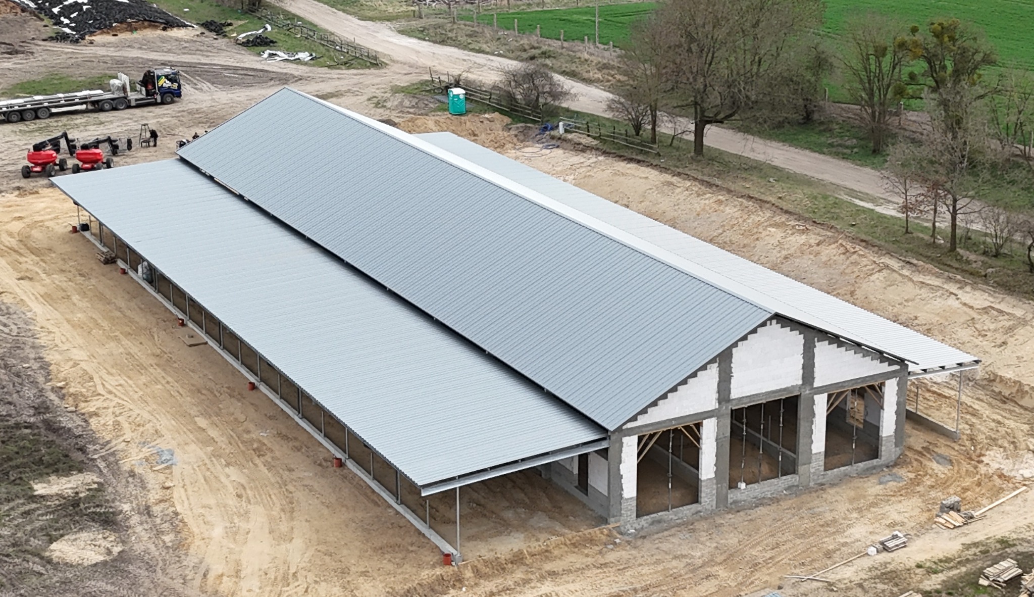 Budowa hali z szarej blachy falistej, widok z góry. Częściowo wykończone ściany, teren budowy, sprzęt budowlany w tle. Dwa segmenty hali.