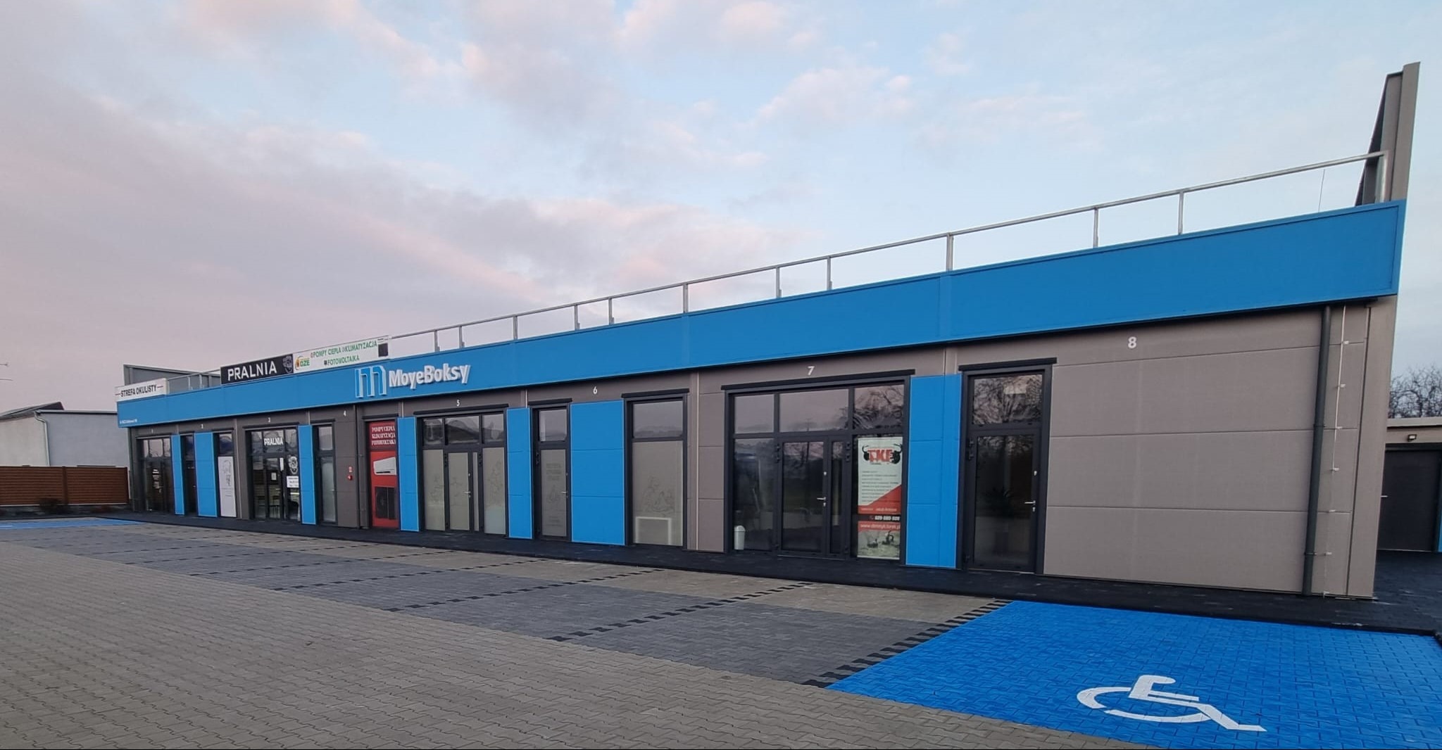 Nowoczesny budynek modułowy z niebieskimi akcentami i parkingiem, widok z zewnątrz. Na budynku widoczne szyldy różnych firm, np. pralni i 'Moye Boksy'.