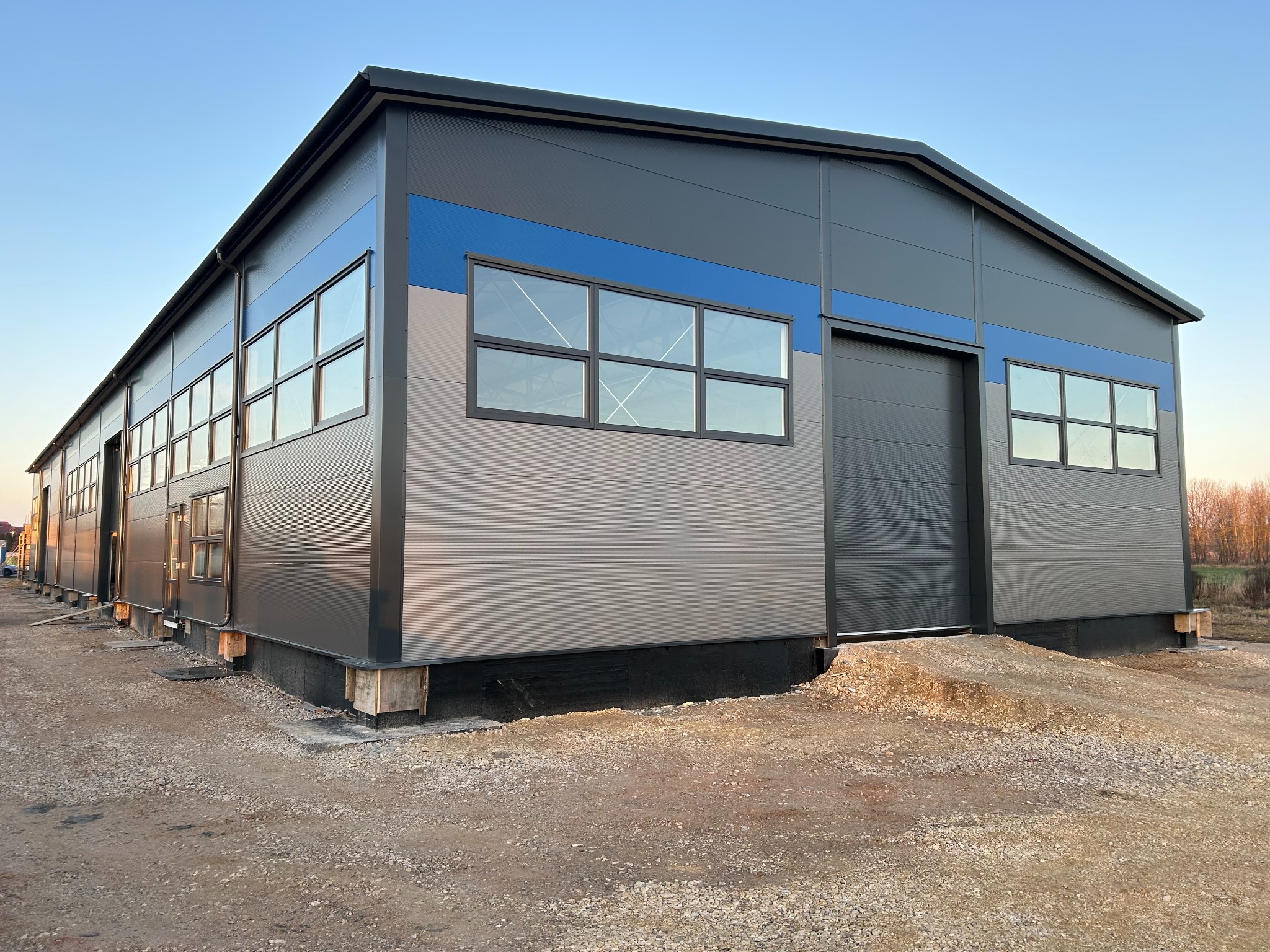 Nowoczesna hala stalowa w szaro-niebieskiej kolorystyce, z widocznymi oknami i bramą segmentową, na placu budowy z gruzem. Ujęcie z dołu, wczesny wieczór.