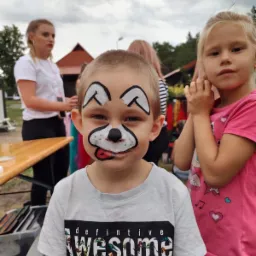 Chłopiec z pomalowaną twarzą w motywie psa, widoczna wystawiona część języka, na zewnątrz, w tle inne dzieci i osoby dorosłe.