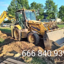 Żółta koparko-ładowarka Komatsu na placu budowy, z widocznym wykopem i numerem telefonu na szybie, w tle domy i zieleń.