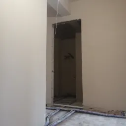 Surowe, jasne wnętrze w trakcie prac wykończeniowych, z widocznymi przewodami instalacyjnymi na podłodze i gładkimi ścianami w odcieniach beżu, z otwartym przejściem do kolejnego pomieszczenia.