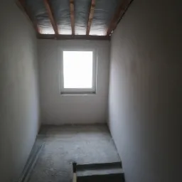 Surowe wnętrze klatki schodowej w trakcie prac wykończeniowych, z widocznym oknem, surowymi ścianami po nałożeniu tynku i drewnianą konstrukcją stropu.