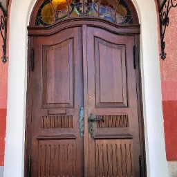 Dwuskrzydłowe, ciemnobrązowe, drewniane drzwi wejściowe z dekoracyjnym, półokrągłym witrażem nad nimi, umieszczone w budynku z białymi i czerwonymi elementami elewacji.