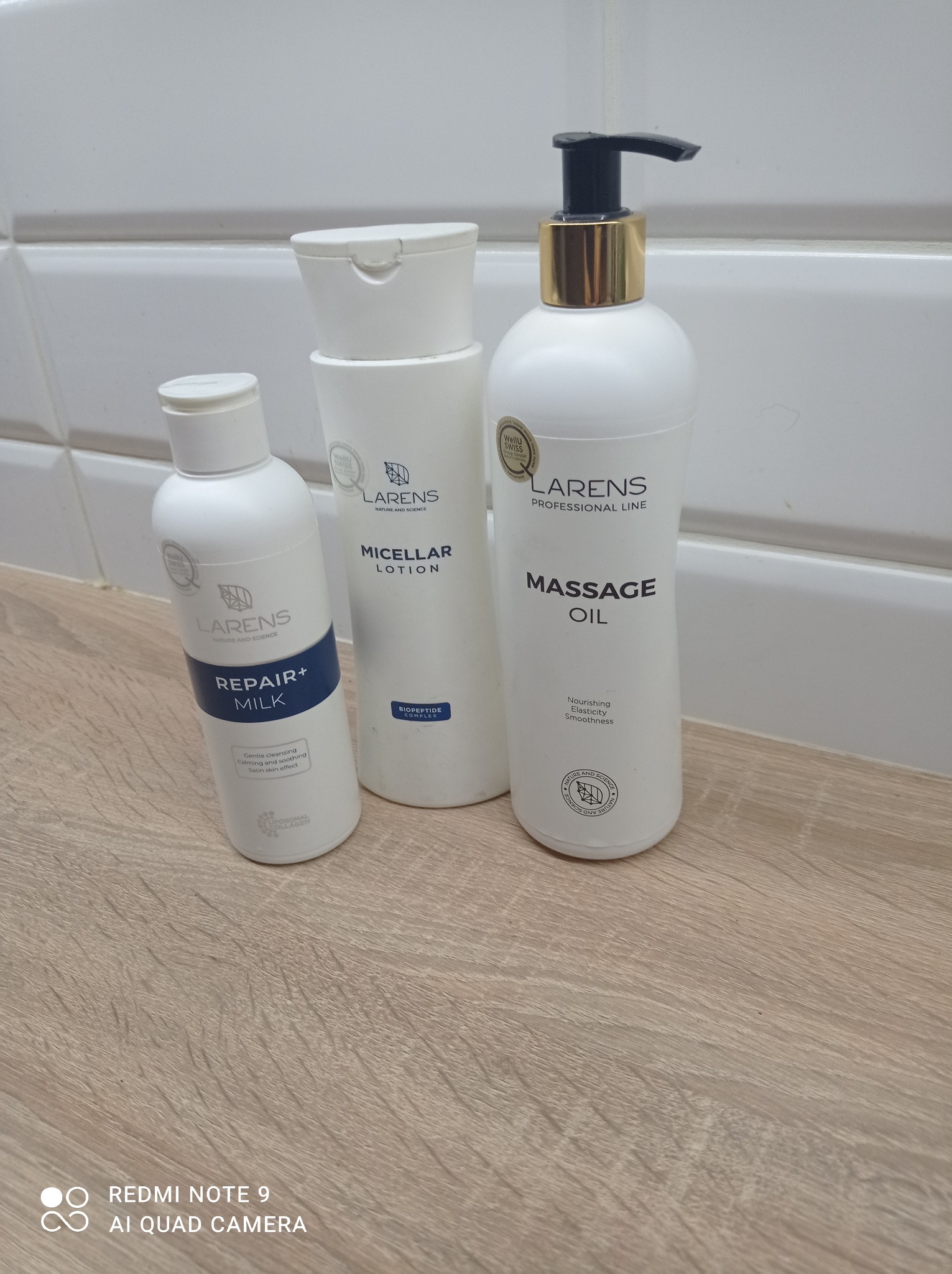Trzy białe butelki kosmetyków marki Larens: mleczko do demakijażu, lotion micelarny i olejek do masażu, ustawione obok siebie na blacie z imitacją jasnego drewna na tle białych płytek ściennych.