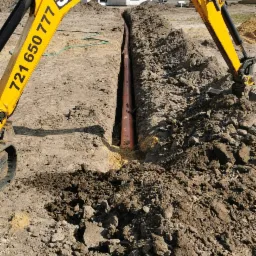 Żółta koparka JCB z numerem telefonu na ramieniu nad wykopanym rowem z pomarańczową rurą, na tle domu w budowie i błękitnego nieba.