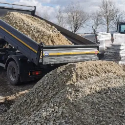Ciężarówka z podniesioną skrzynią wyładowuje materiał sypki, mieszankę piasku i kamieni, na placu budowy. W tle widoczne są palety z materiałami budowlanymi oraz mała koparka.