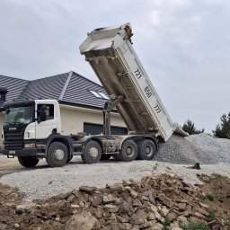 Adrian Lejczak - Ciężarówka Scania wyładowuje żwir na placu budowy przed domem z ciemną dachówką. Widoczny stos żwiru i gruz. Pochmurne niebo w tle.