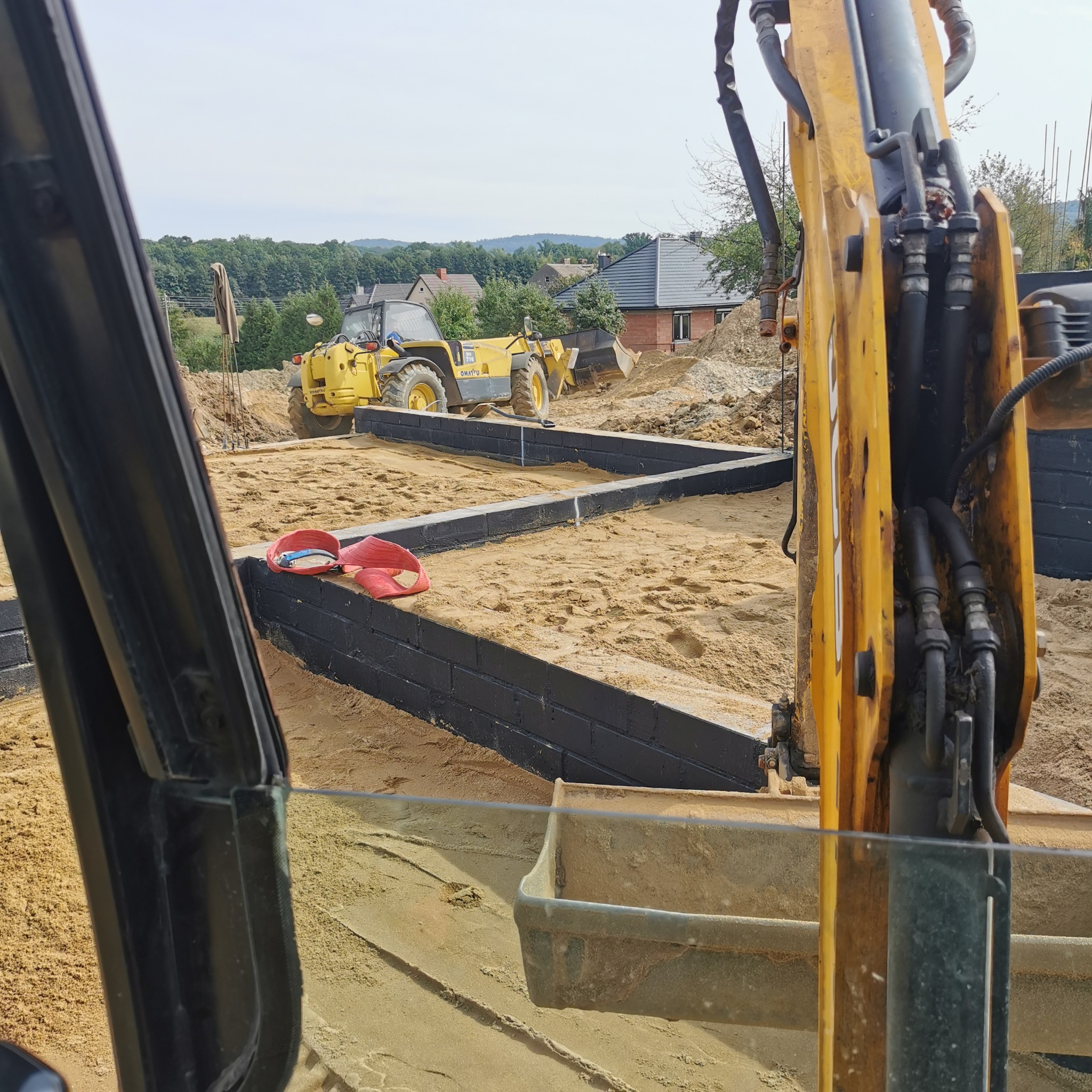 Przygotowanie terenu pod fundamenty: widok z kabiny koparki na wypełniony piaskiem wykop z czarnymi bloczkami, w tle żółta ładowarka i zabudowa jednorodzinna.