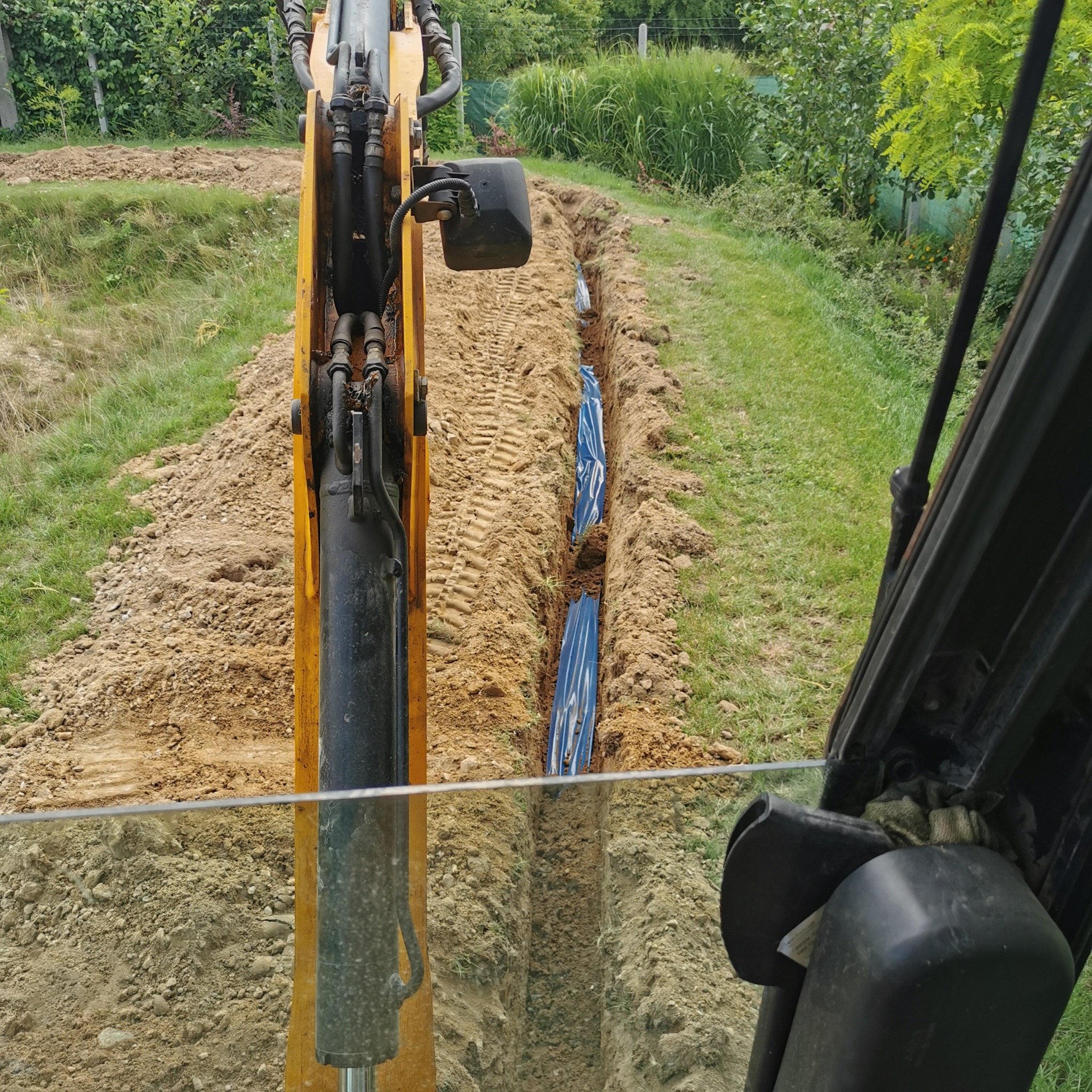 Widok z kabiny koparki na wykopany rów z ułożoną niebieską rurą drenażową, ziemia po bokach, trawa i zieleń w tle.