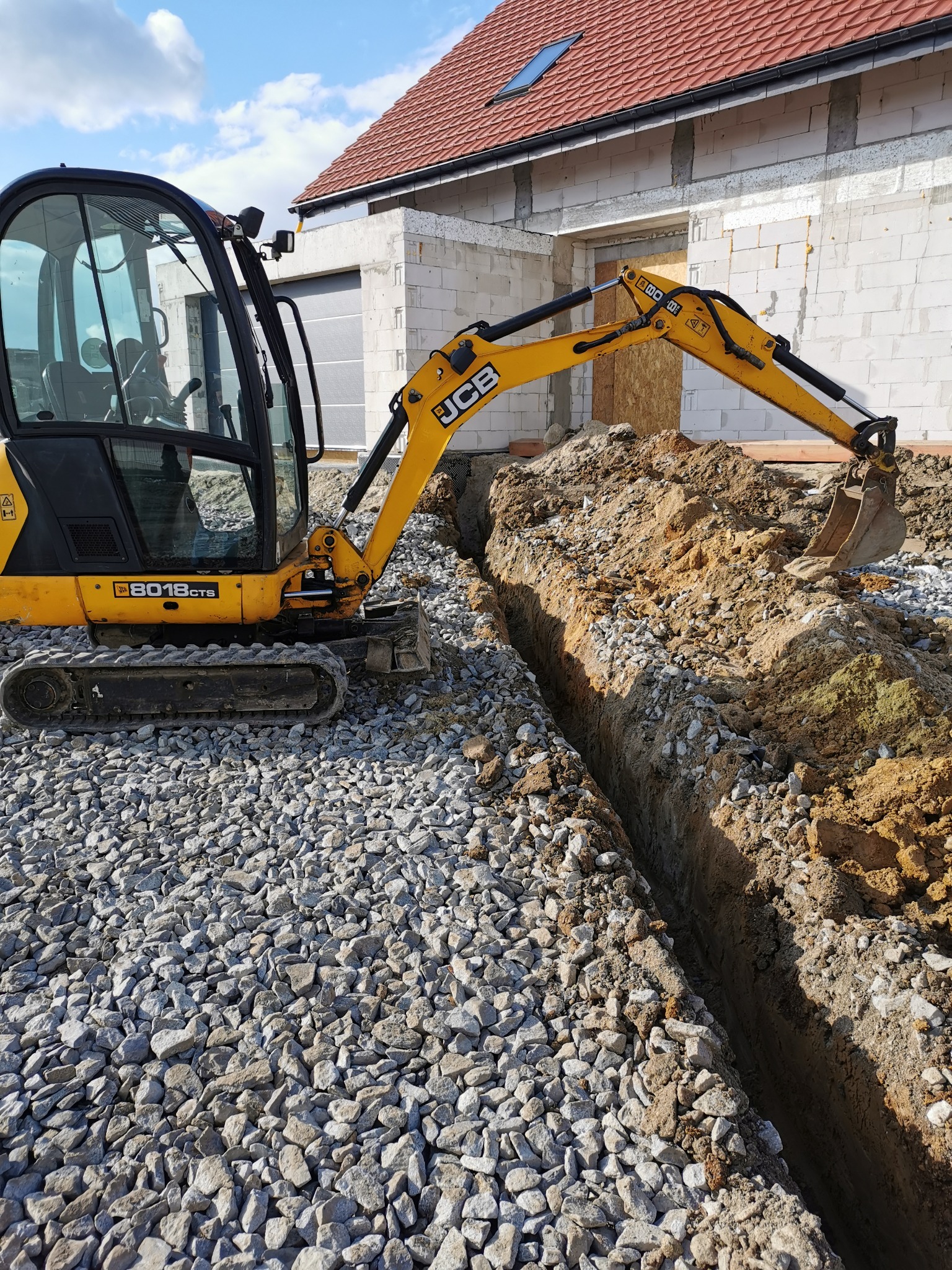 Żółta koparka JCB 8018 CTS wykonuje wąski wykop w ziemi, na tle wznoszonego budynku z czerwonym dachem i szarą bramą garażową.