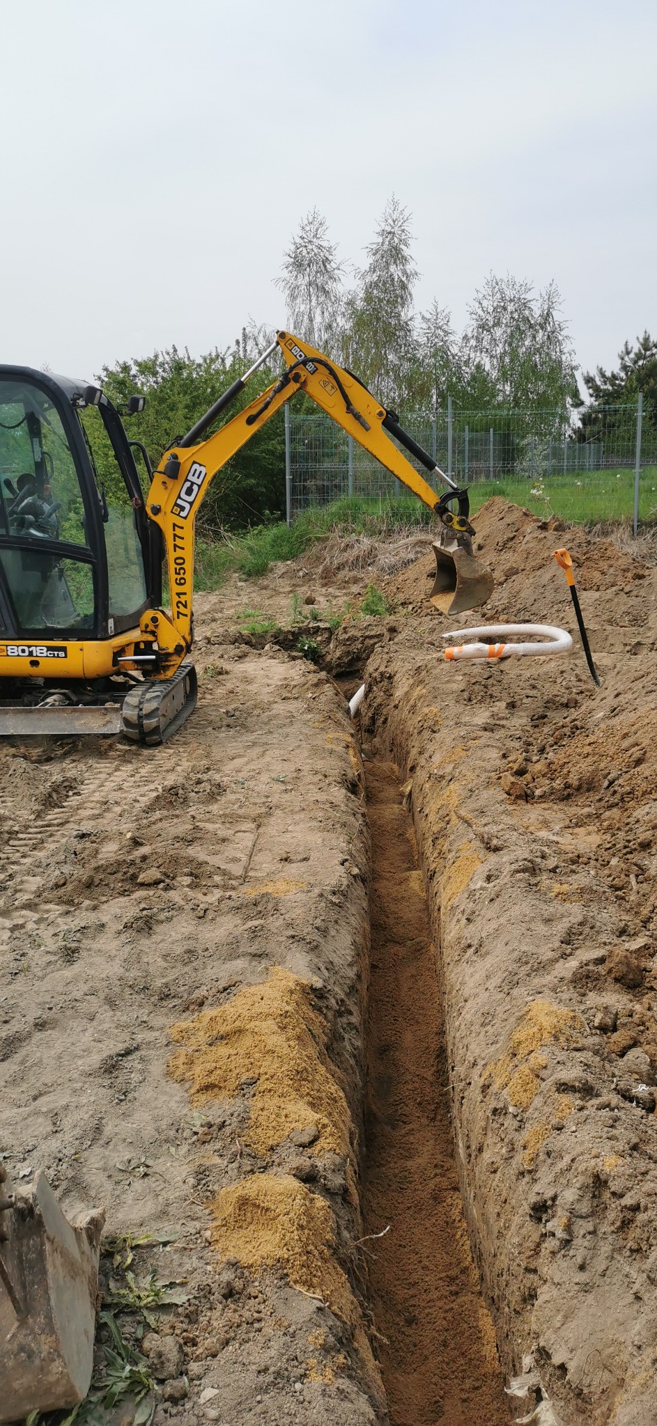 Żółta mini koparka JCB przygotowuje wąski wykop pod rury drenażowe, widoczna ułożona rura karbowana i szpadel.