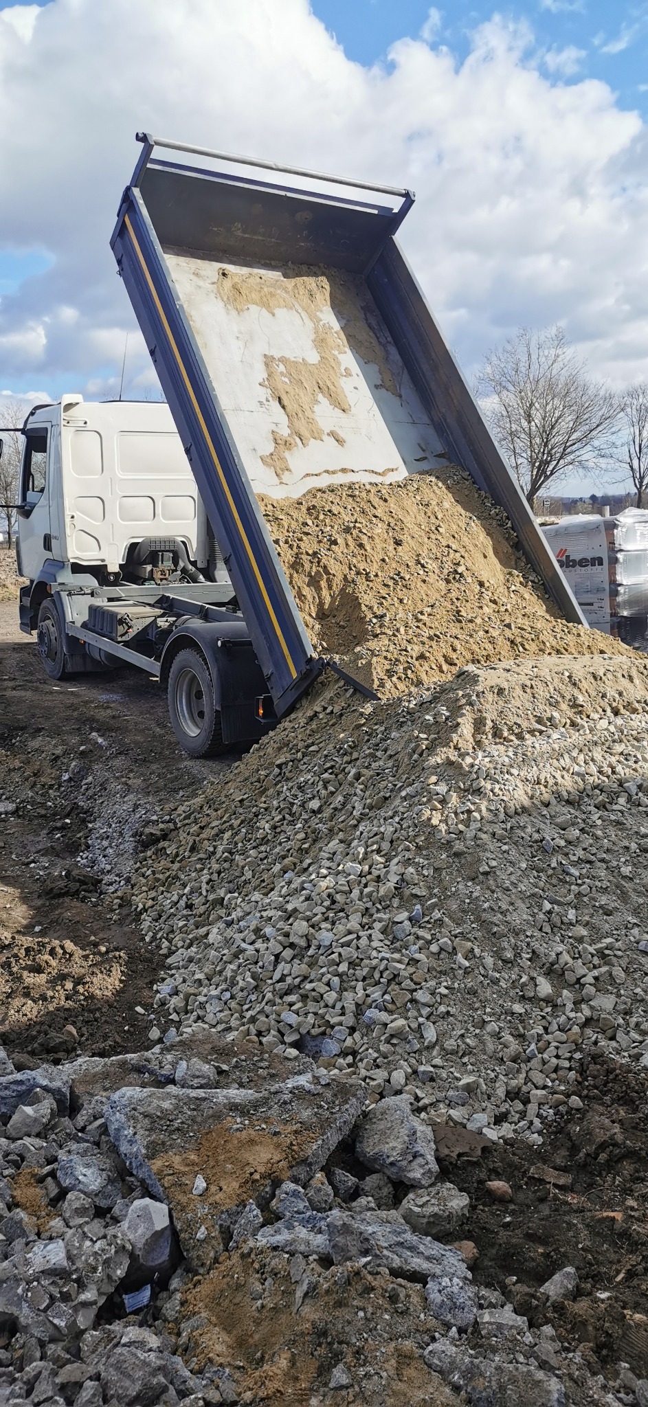 Ciężarówka z podniesioną skrzynią wyładowuje mieszankę piasku i żwiru na ziemię, tworząc kopiec na placu budowy.