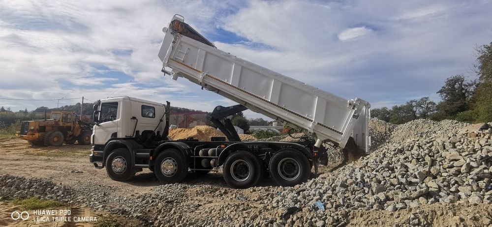 Wyładowywanie kruszywa z białej wywrotki na placu budowy, w tle widoczna żółta ładowarka kołowa Volvo.