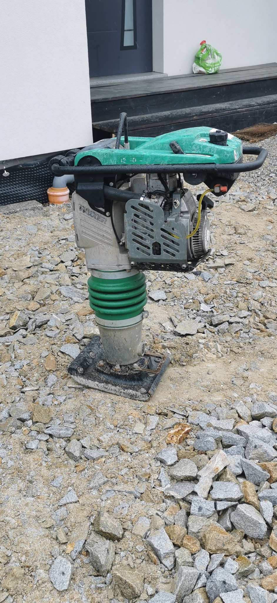 Zielona ubijarka gruntu marki Wacker stoi na warstwie kamieni i piasku przed budynkiem z czarnymi drzwiami.