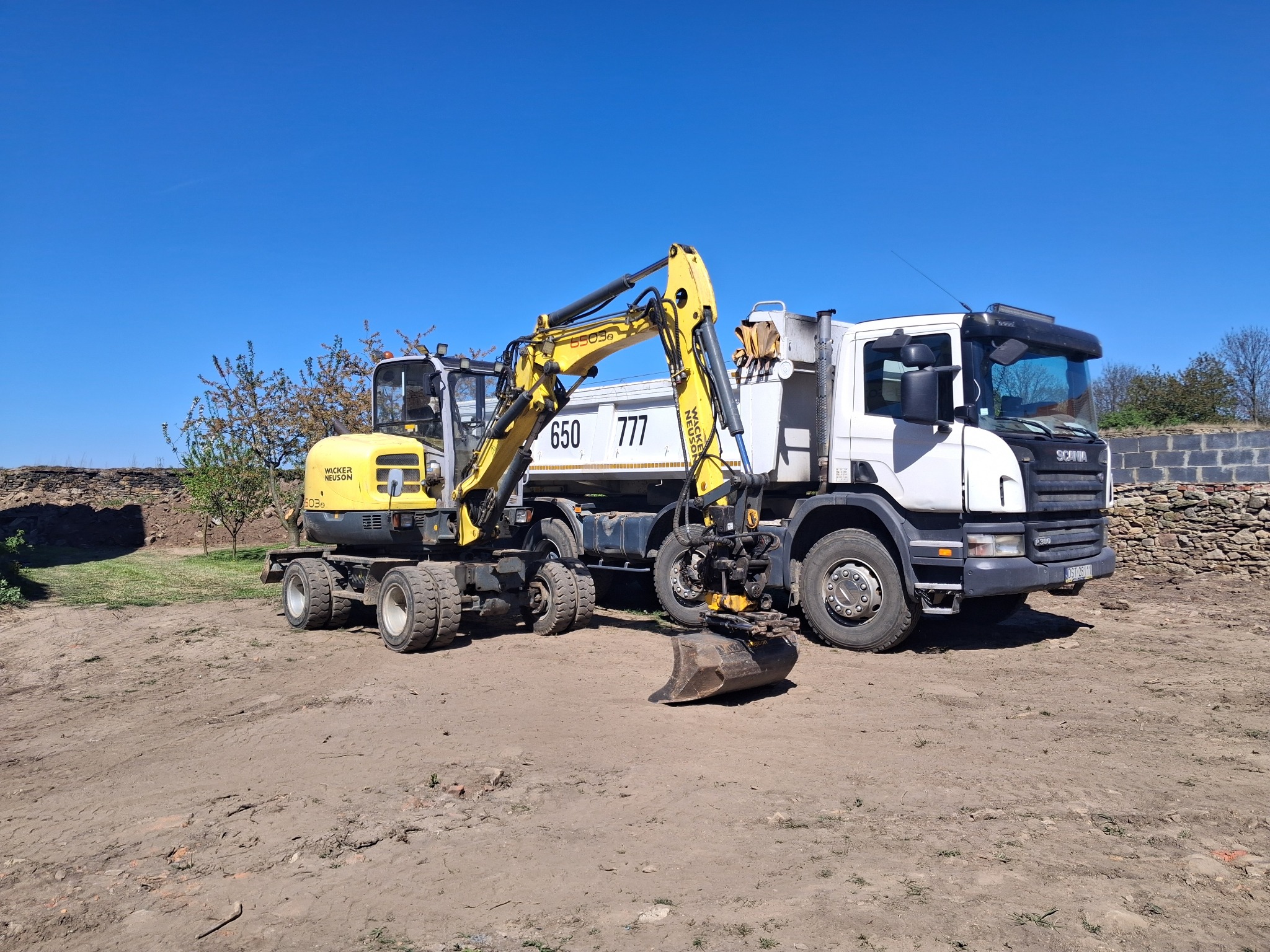 Żółta koparka Wacker Neuson i biała ciężarówka Scania na placu budowy w słoneczny dzień. Koparka z łyżką opuszczoną na ziemię, gotowa do pracy.