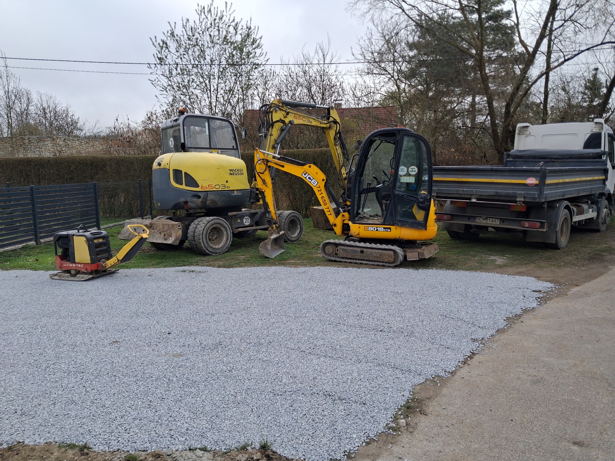 Utwardzony teren żwirowy z użyciem zagęszczarki i dwóch koparek JCB, obok wywrotka. Widok na posesję w pochmurny dzień.