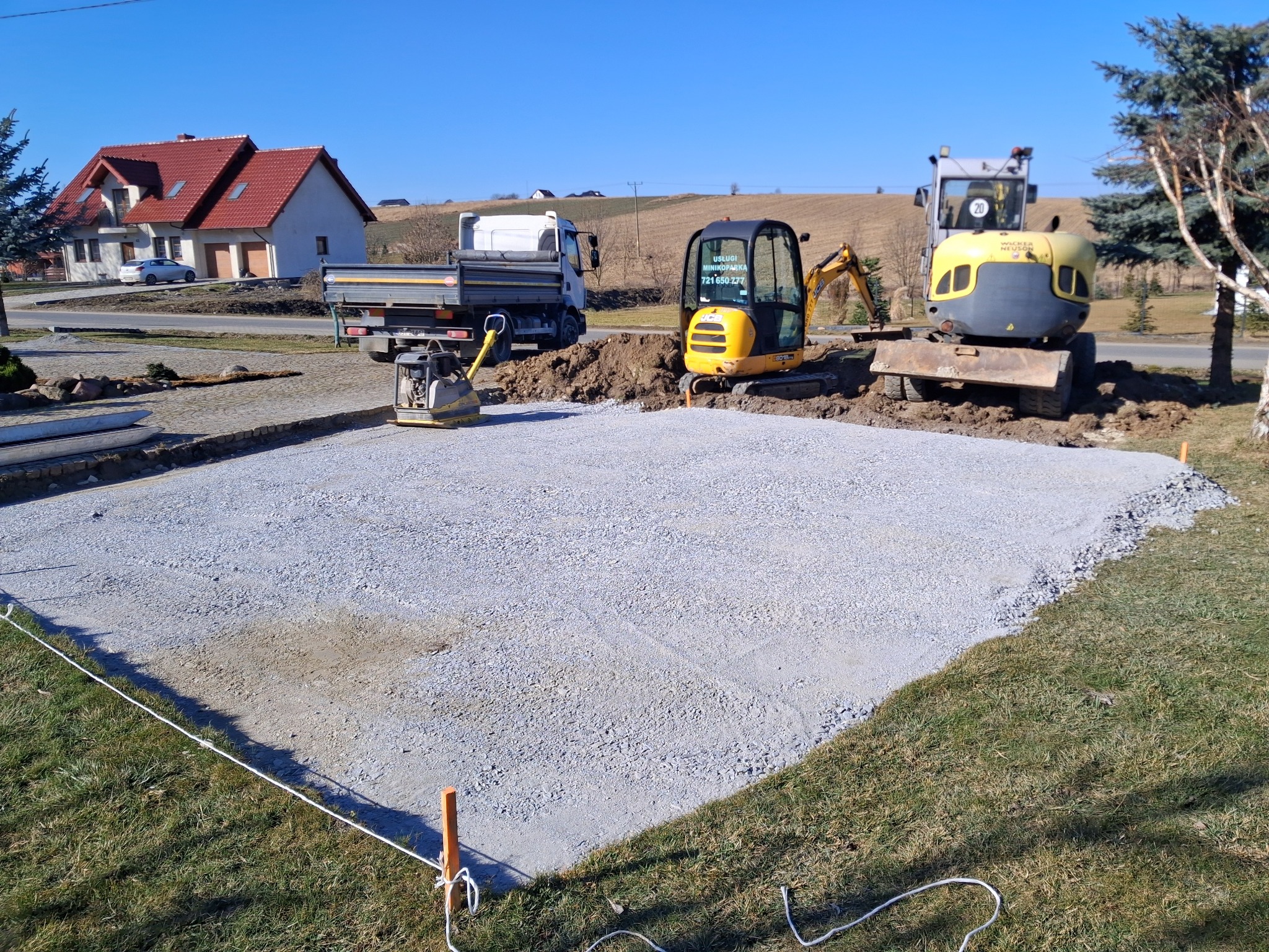 Przygotowany teren pod budowę z użyciem mini koparek i ubijarki. Widoczny wyznaczony obszar z równo rozłożonym żwirem, ciężarówka i dom w tle.