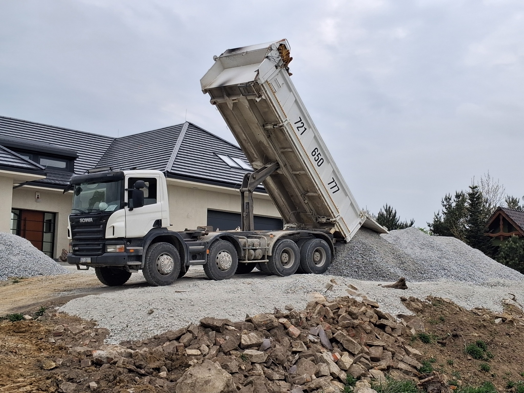 Ciężarówka Scania wyładowuje żwir na placu budowy przed domem z ciemną dachówką. Widoczny stos żwiru i gruz. Pochmurne niebo w tle.
