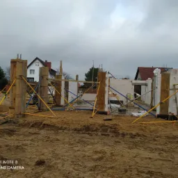 Wznoszenie ścian domu murowanego z betonu komórkowego, widoczne szalunki i podpory stabilizujące, w tle zabudowa jednorodzinna i pochmurne niebo.