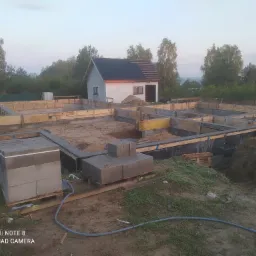 Wykopane fundamenty z szalunkiem drewnianym i izolacją, w tle budynek gospodarczy z częściowo położoną dachówką.