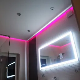 Nowoczesna łazienka z podświetlanym lustrem, różowym oświetleniem LED, umywalką nablatową i szarą ścianą.