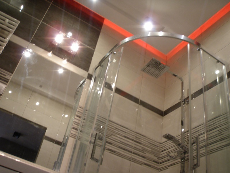 Nowoczesna kabina prysznicowa z chromowanymi elementami, oświetleniem LED w kolorze czerwonym i lustrem z odbiciem płytek w odcieniach szarości i bieli.