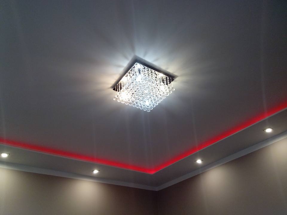 Kryształowy żyrandol sufitowy z podświetleniem LED w kolorze czerwonym, zainstalowany w narożniku pomieszczenia z szarą ścianą.