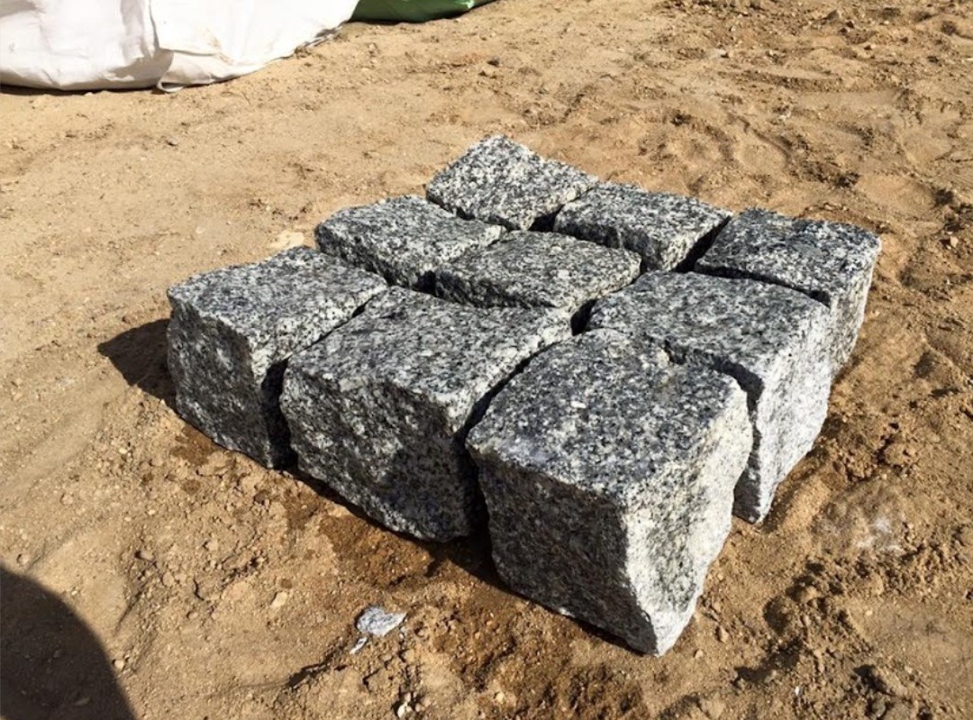 Granitowa kostka brukowa ułożona w kwadrat na piaszczystym podłożu, widoczny worek z materiałem budowlanym w tle.