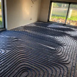 Instalacja ogrzewania podłogowego z rur w kolorze niebieskim, rozłożona na folii z siatką w surowym wnętrzu z dużymi oknami, widoczne cegły i zieleń za oknem.