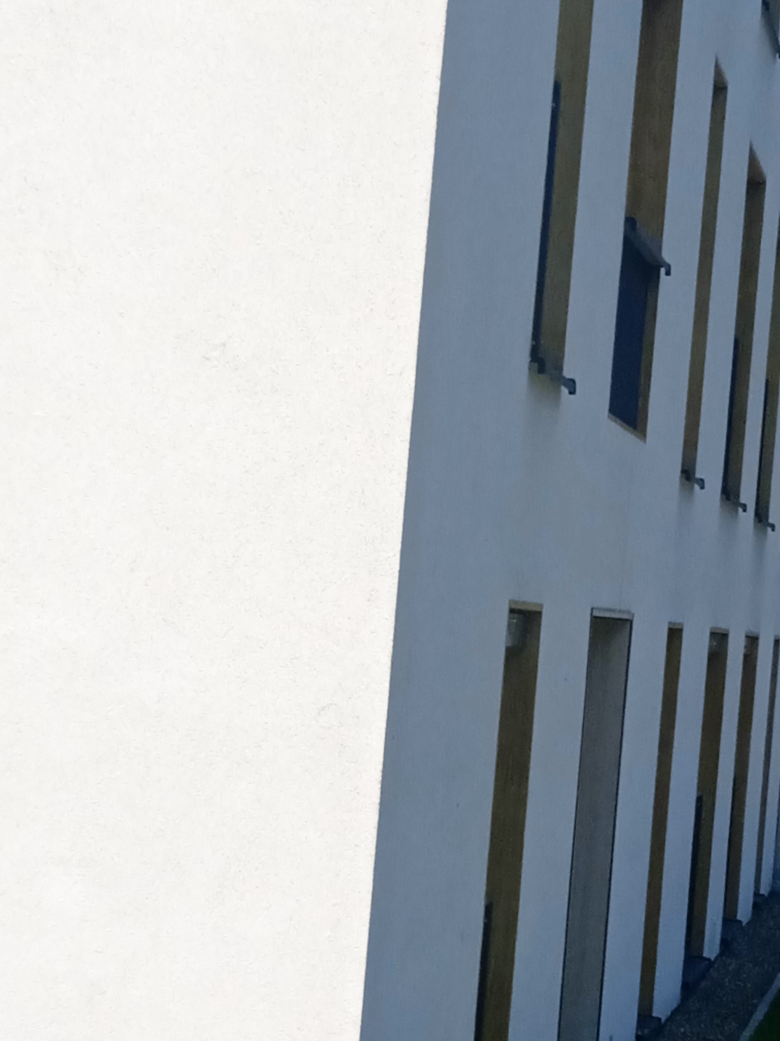 Fragment białej elewacji budynku z pionowymi wnękami i oknami, widzianej pod kątem, z wyraźnym cieniem na gładkiej powierzchni.
